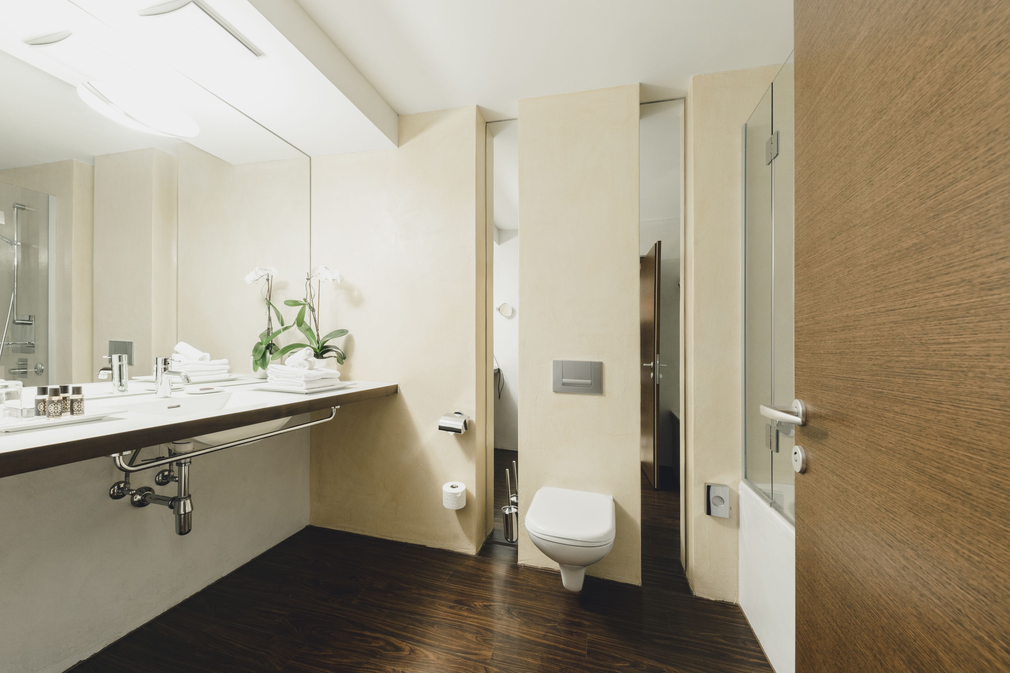 A modern bathroom with a wooden floor, a white sink with a towel rack, a toilet, and a mirror.