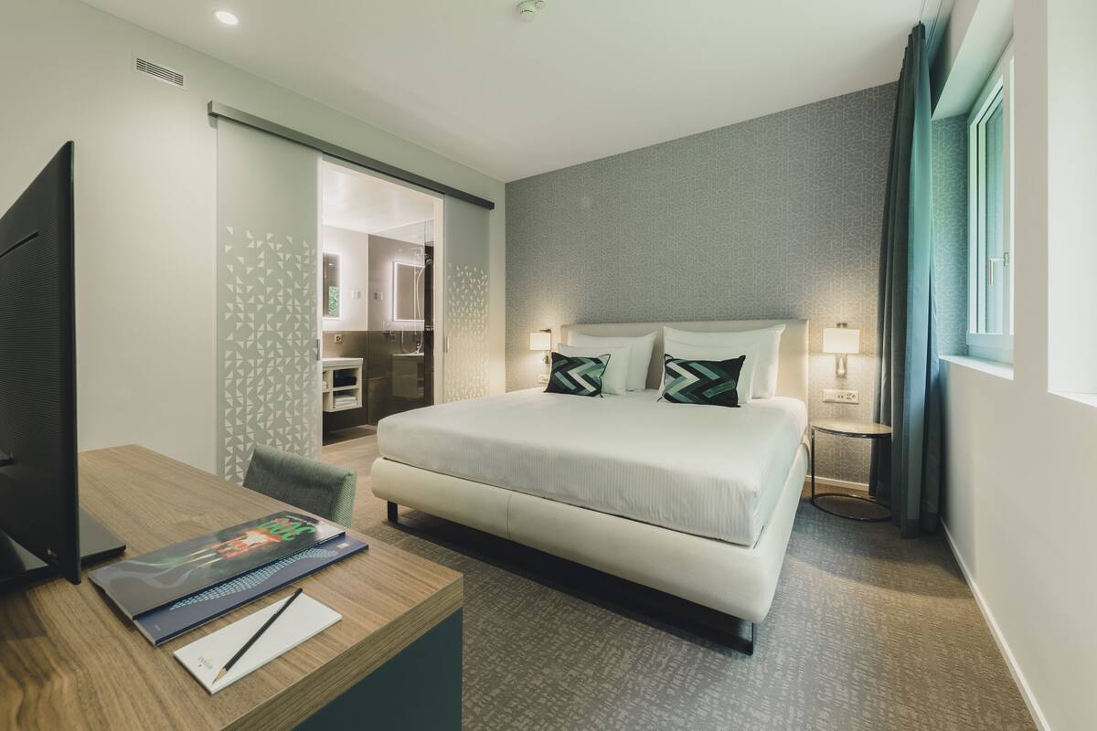 Habitación de hotel con cama, escritorio con computadora, televisor y baño visible a través de una puerta corrediza.