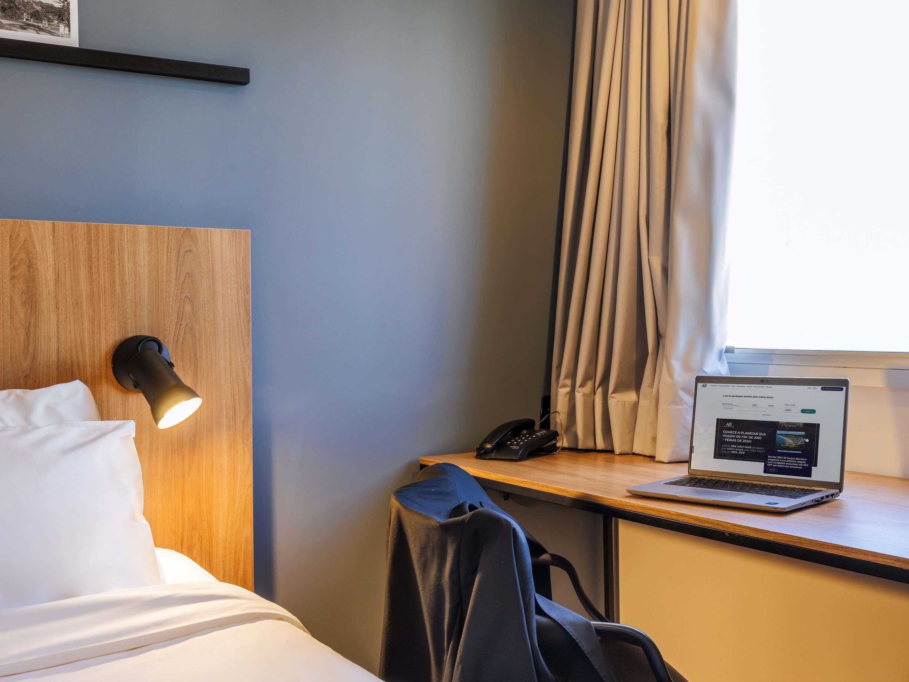 A hotel room with a bed, a desk with a laptop, a phone, and a wall-mounted light.