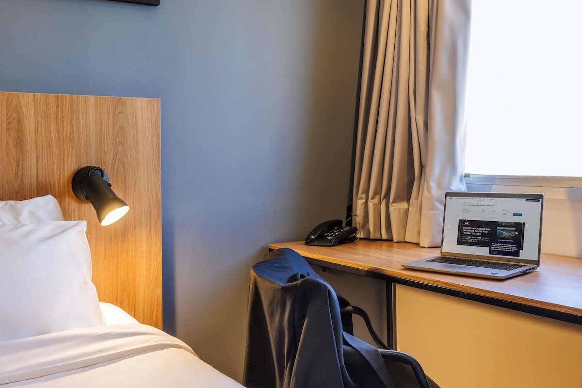 A hotel room with a bed, a desk with a laptop, a phone, and a wall-mounted light.