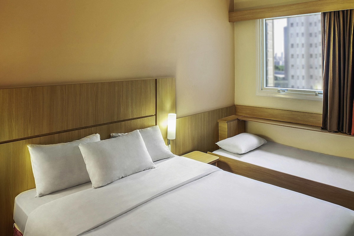Une chambre d’hôtel avec un grand lit, un lit plus petit, une table de chevet, une lampe et une fenêtre avec des rideaux.