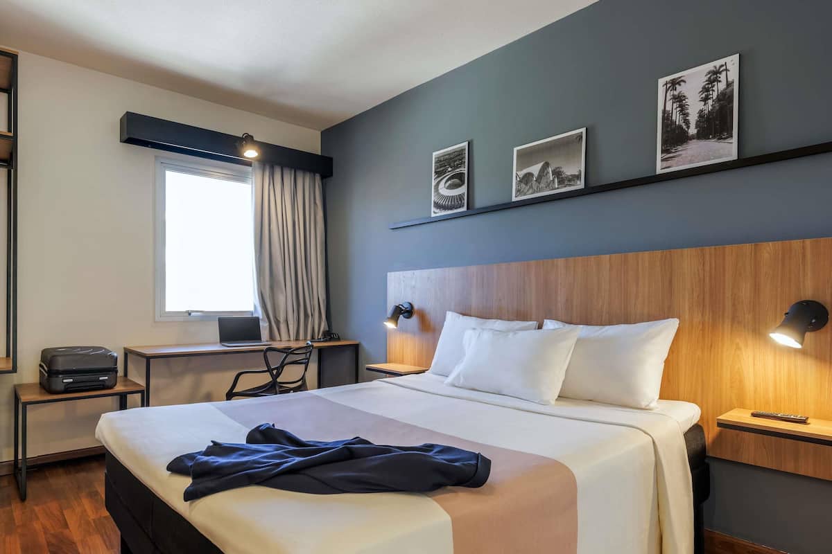 Quarto de hotel com cama, escrivaninha e janela com cortinas.