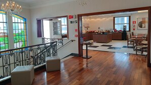 A hotel lobby with a wooden floor, a staircase with a wrought iron railing, a chandelier, and a view of a garden through a stained glass window. - Ibis Belo Horizonte Liberdade (Belo Horizonte)