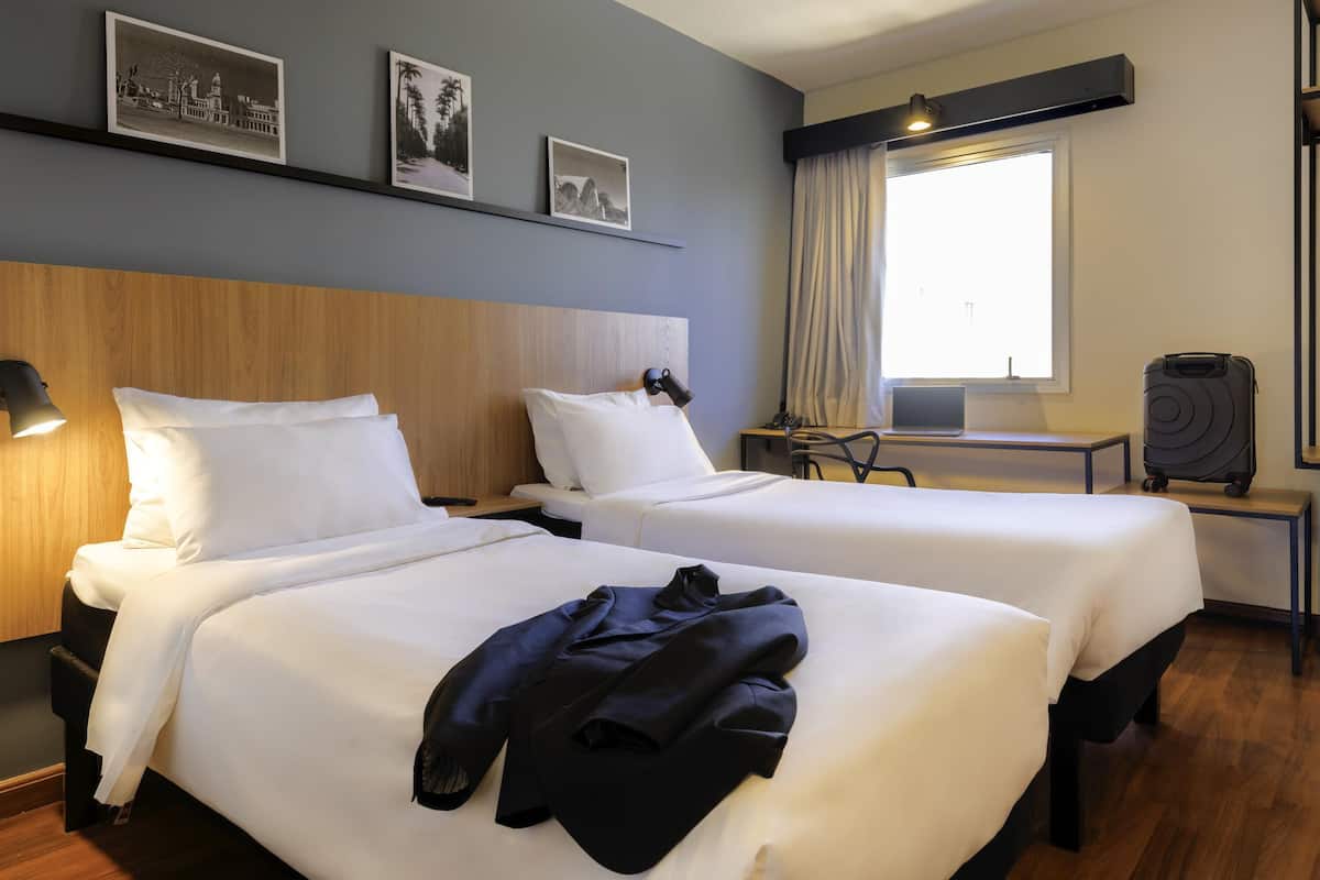 Une chambre d’hôtel avec deux lits, un bureau, un ordinateur portable, une valise et des tableaux encadrés au mur.