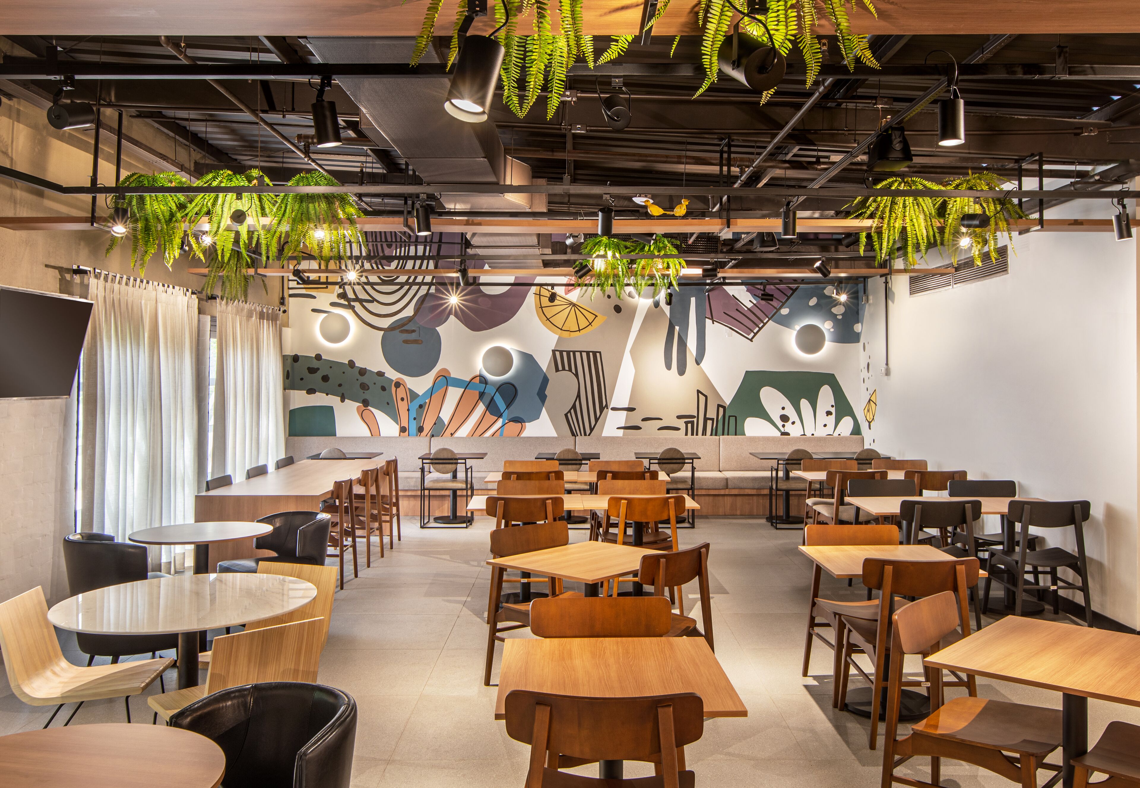 Un café moderno con mesas y sillas de madera, plantas colgantes y un mural colorido en la pared.