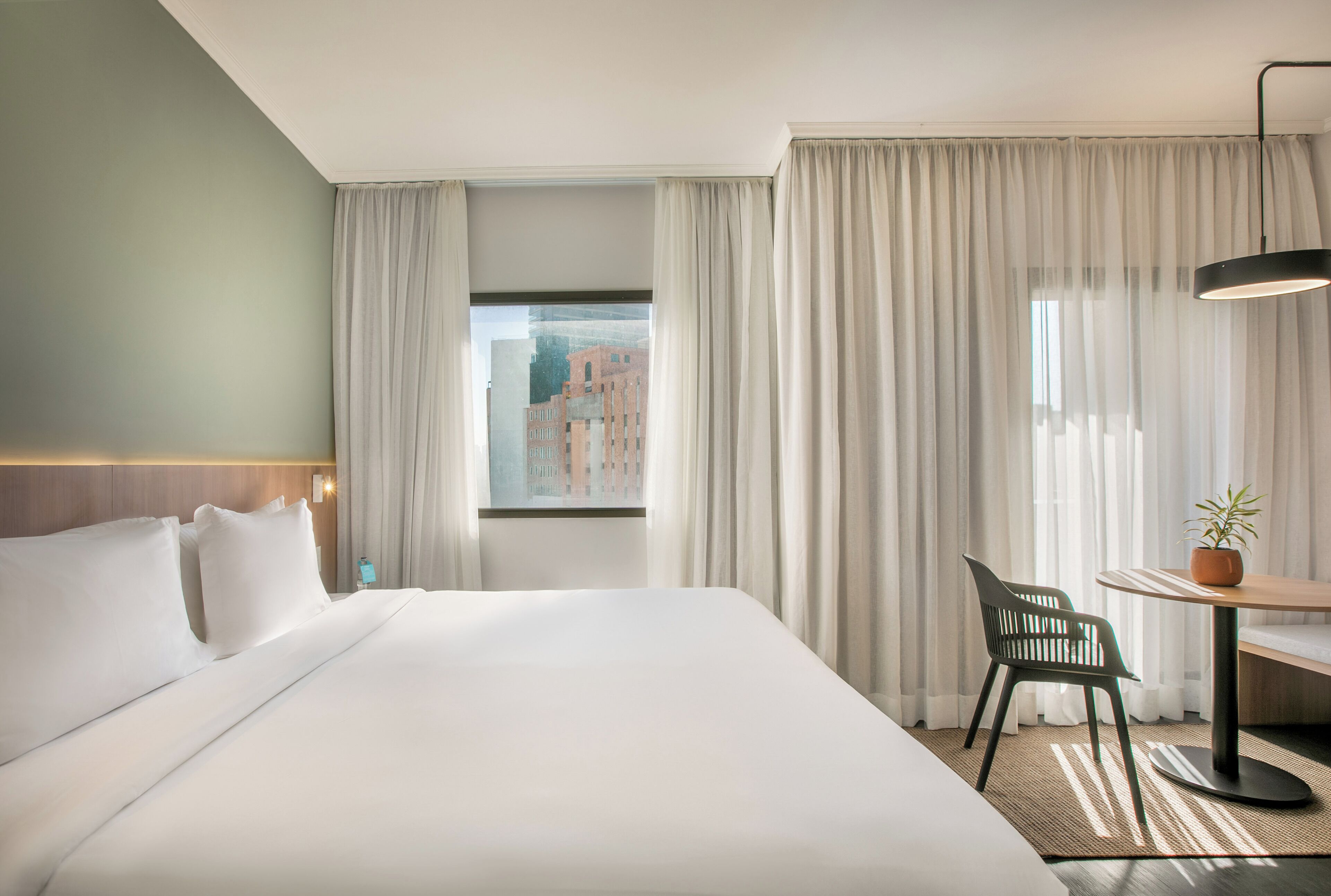 Una habitación de hotel con una cama grande, un escritorio con una silla, una planta en maceta y vistas a edificios a través de cortinas translúcidas.