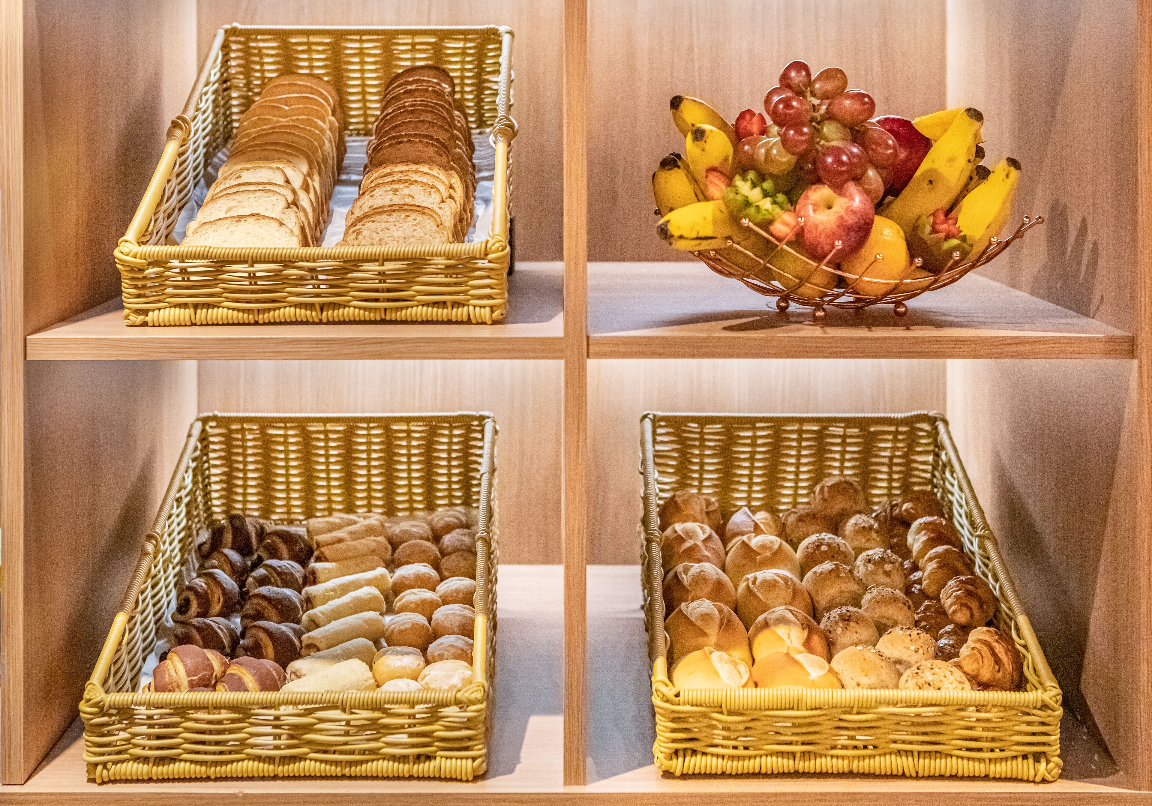 Four wicker baskets on shelves containing bread, pastries, and a fruit bowl.