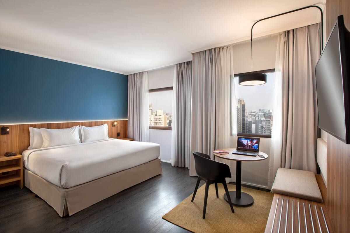 Ein Hotelzimmer mit einem Bett, einem Schreibtisch mit Laptop, einem Fernseher und Blick auf die Stadt.
