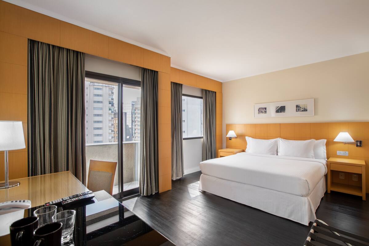 Ein Hotelzimmer mit einem großen Bett, einem Schreibtisch mit Stuhl, einem Fenster mit Vorhängen und Blick auf Gebäude.