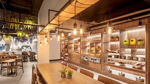 Un restaurante con mesas y sillas de madera, plantas colgantes y estantes con cristalería y productos alimenticios.