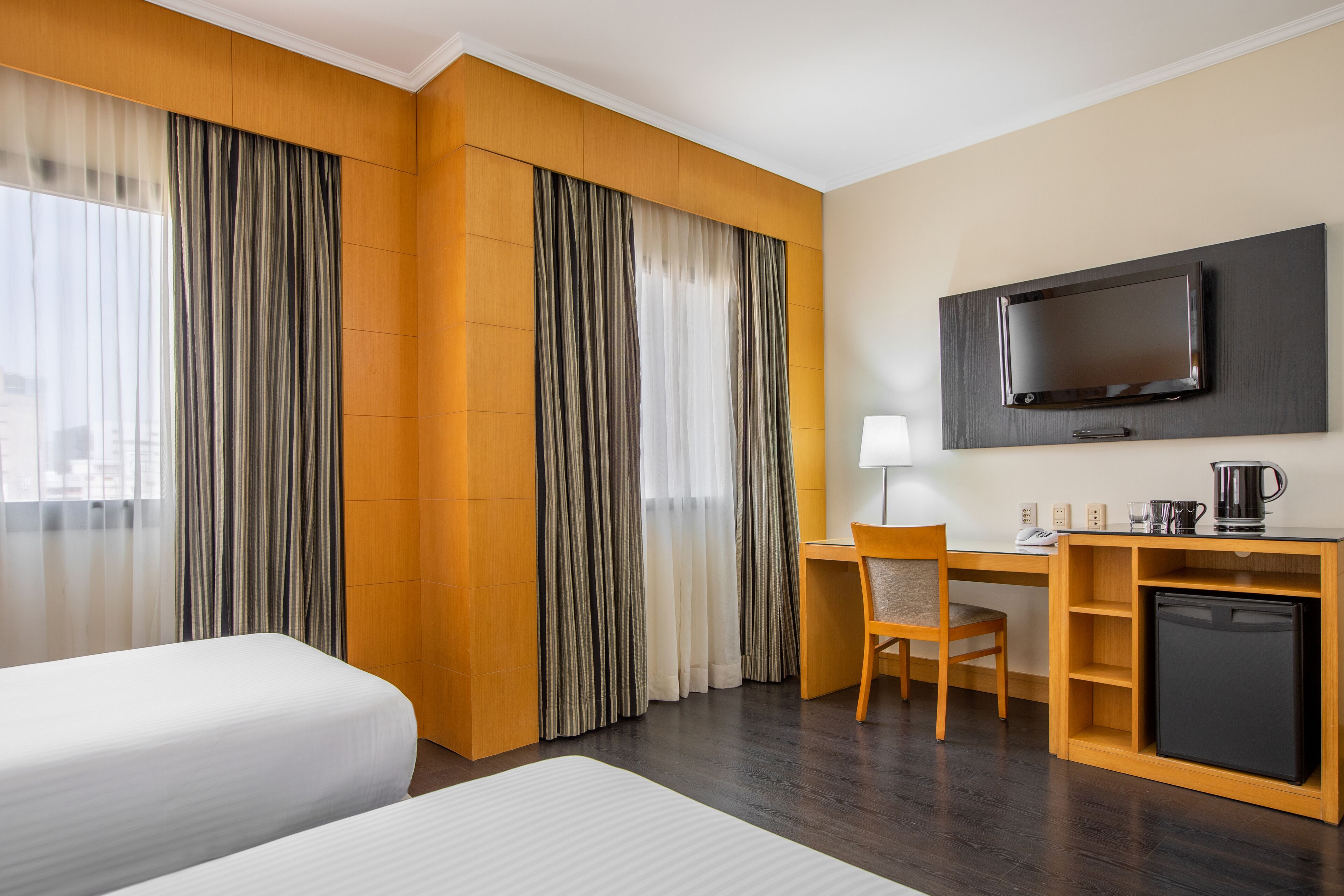 A hotel room with two beds, a desk with a chair, a television mounted on the wall, and a small refrigerator.