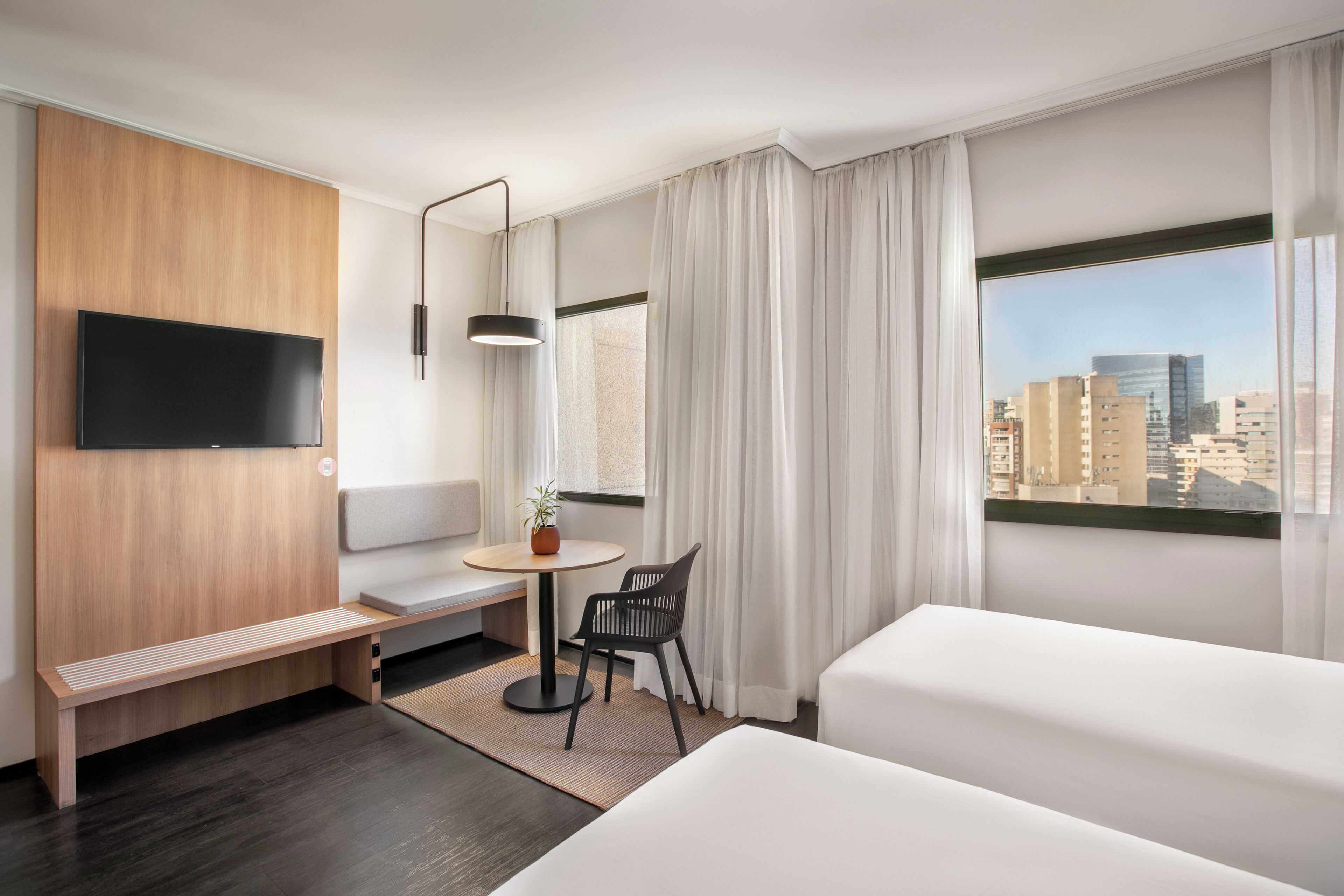 A hotel room with a bed, a desk, a chair, a TV, and a window with a city view.