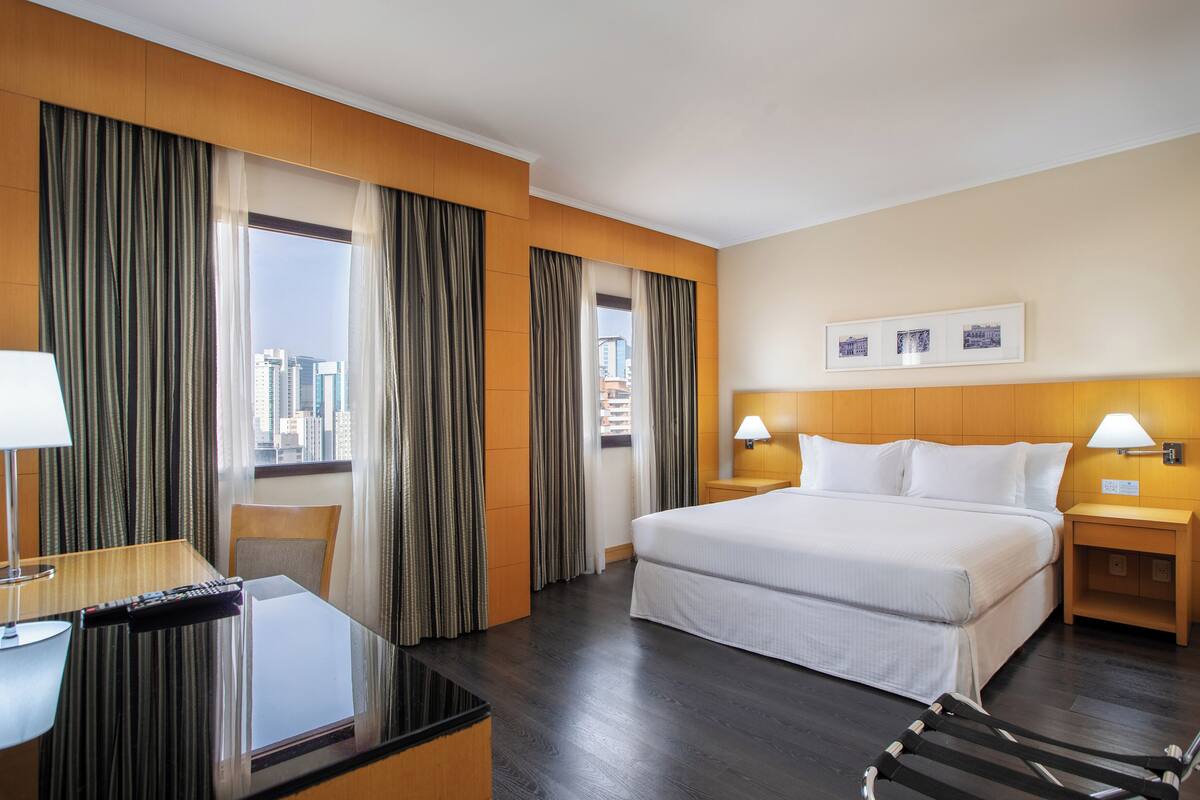 Ein Hotelzimmer mit einem Bett, zwei Nachttischen mit Lampen, einem Schreibtisch mit Stuhl, einer Fernbedienung und Blick auf die Stadt durch große Fenster.