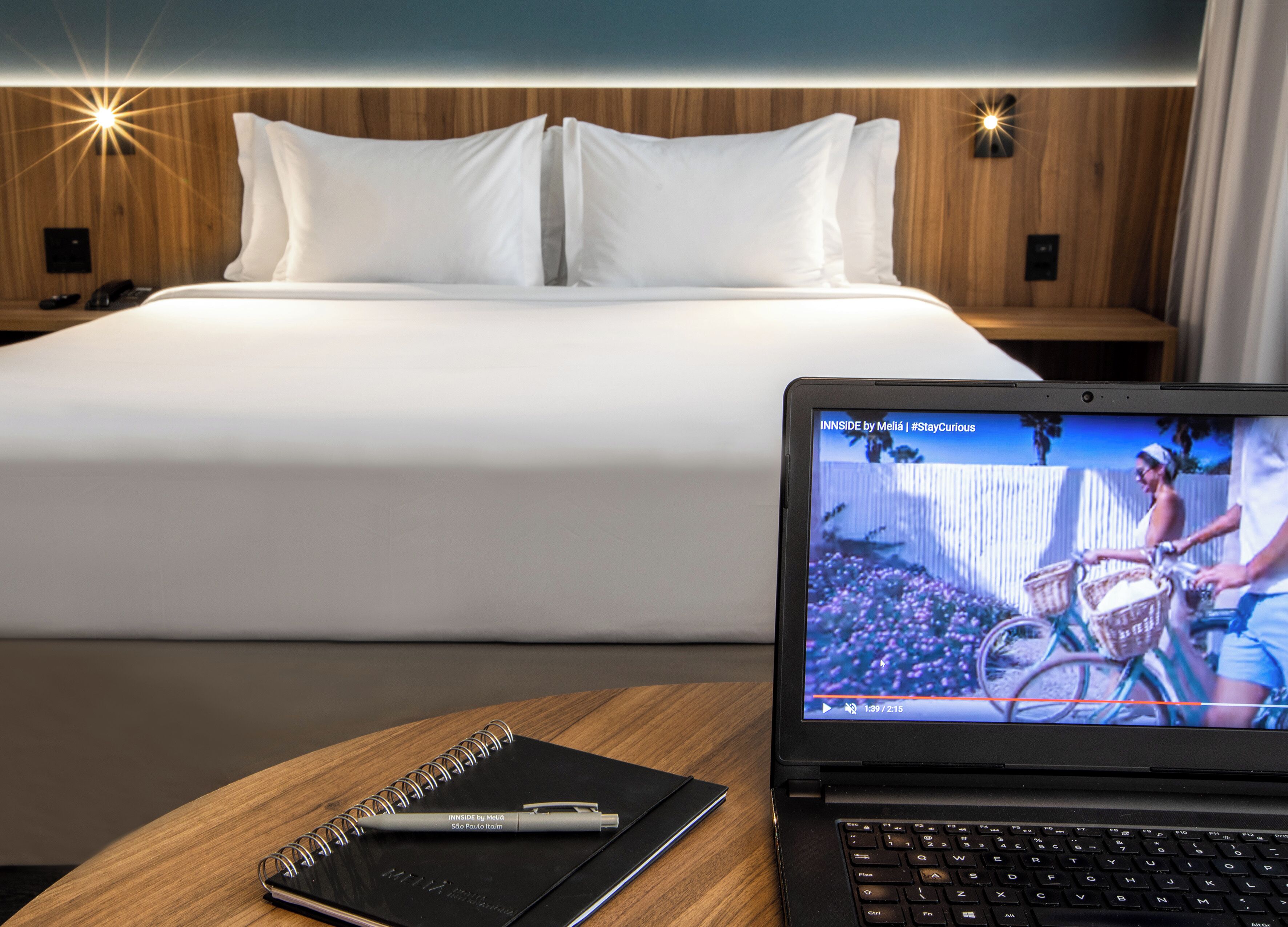 A hotel room with a bed, a laptop on a wooden table, and a notebook with a pen.