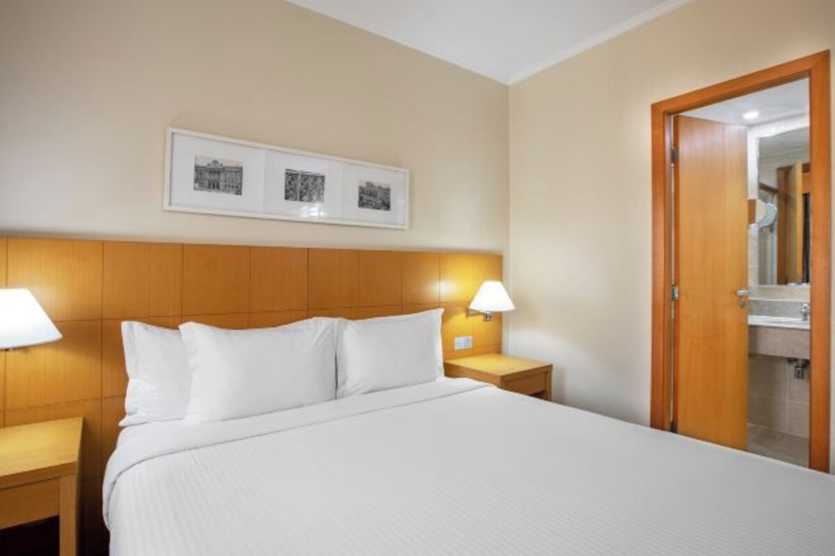 Una habitación de hotel con una cama, dos mesitas de noche con lámparas, un cabecero de madera y una puerta que da a un baño.