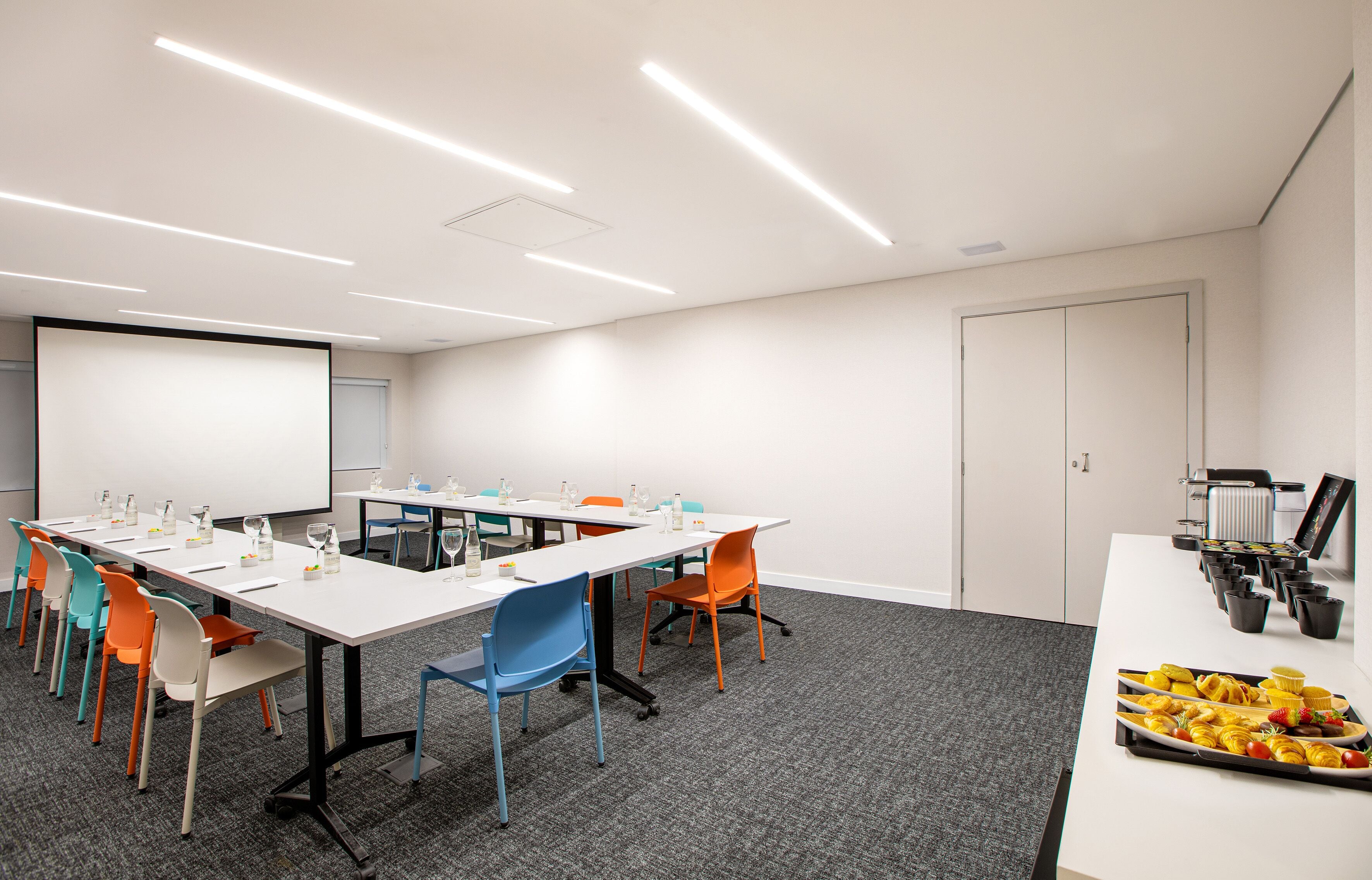 A conference room with white tables, colorful chairs, and a projector screen.