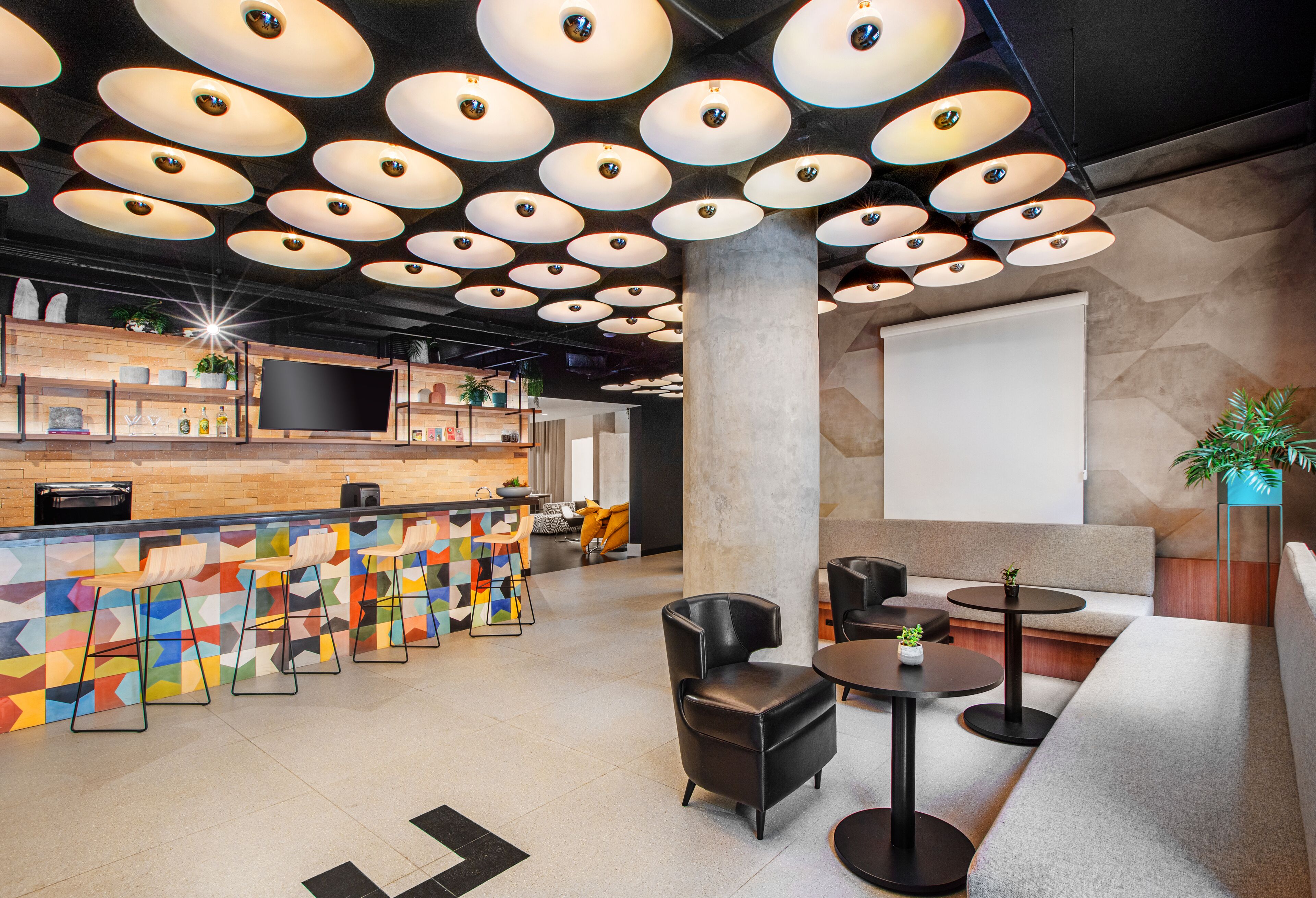Un área de bar moderna con diversas opciones de asientos, incluyendo taburetes y sillas acolchadas, y una pantalla grande para presentaciones.