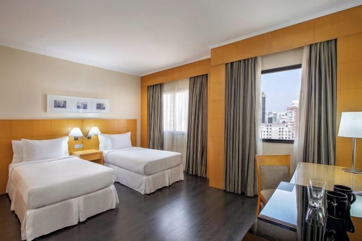 A hotel room with two beds, a desk, a chair, and a view of the city.