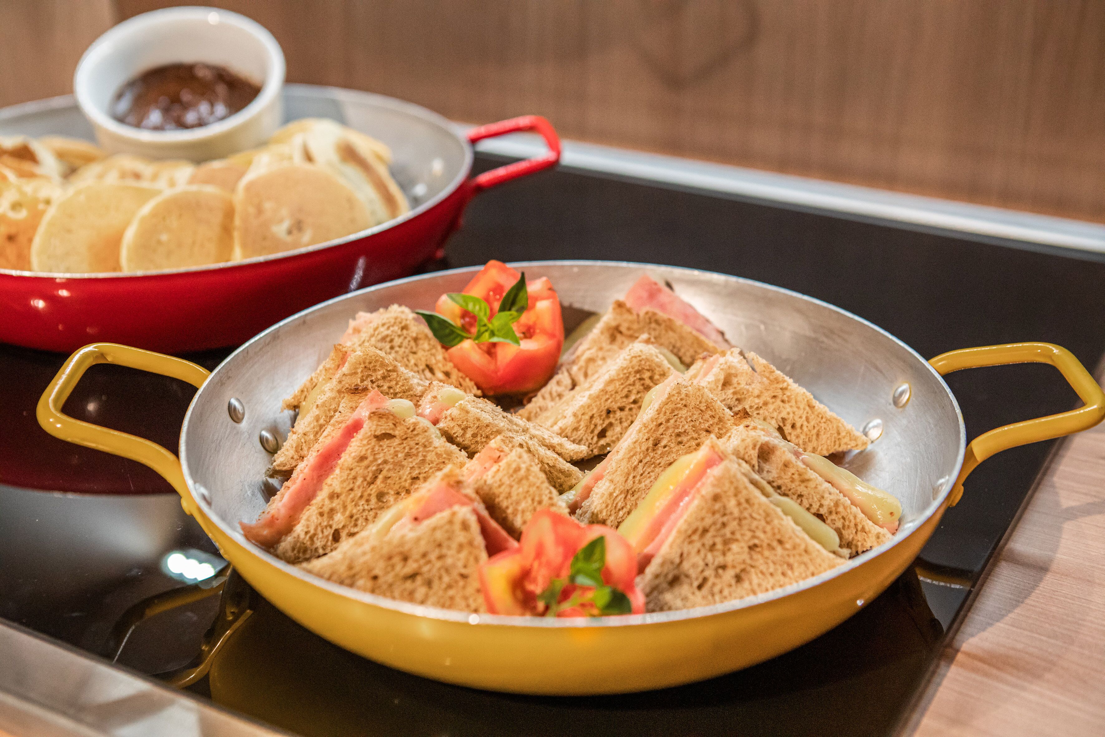 Eine Pfanne mit belegten Sandwiches, Tomaten und Schinken, und im Hintergrund eine rote Pfanne mit weiteren Sandwiches.