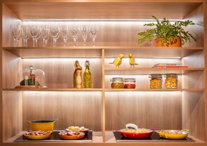 Una cocina con estantes de madera, cristalería, una planta en maceta y diversos alimentos.