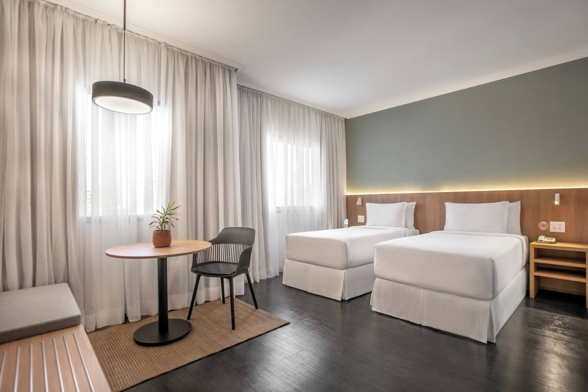 Ein Hotelzimmer mit zwei Einzelbetten, einem kleinen Tisch mit Stuhl, einer Zimmerpflanze und einem großen Fenster mit Vorhängen.