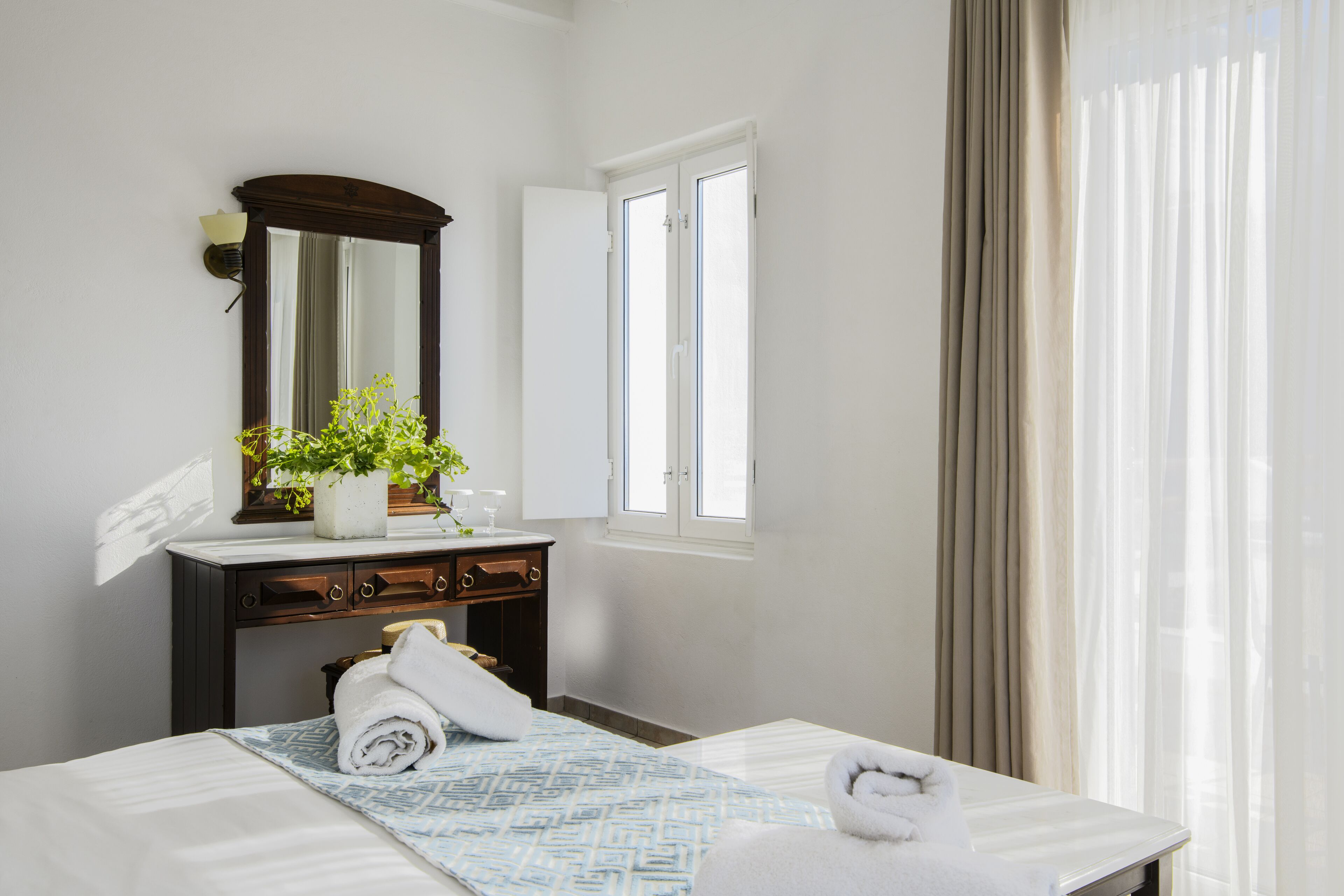 A hotel room with a bed, a dresser, a mirror, a window with curtains, and a potted plant.