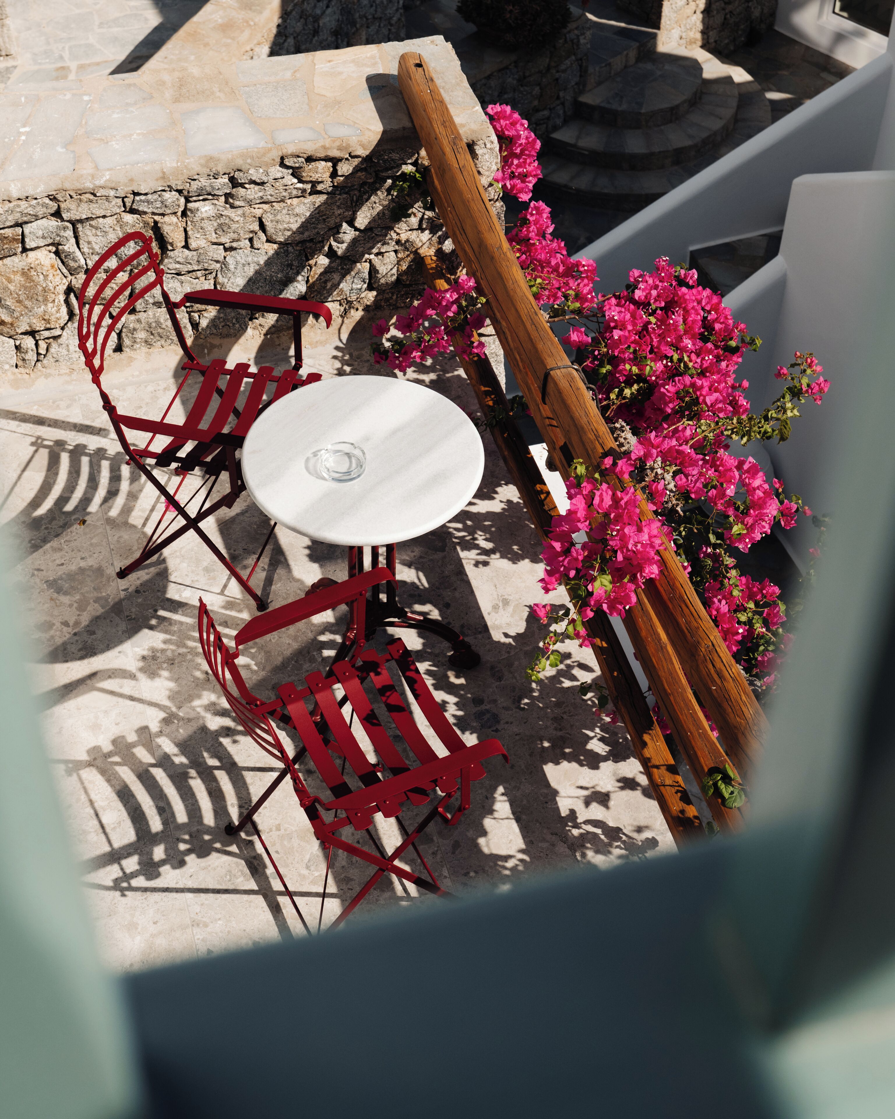 A terrace with a round table and red chairs, surrounded by pink flowers and stone walls.