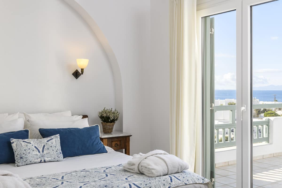 Un dormitorio con cama, almohadas, una mesita de noche, lámpara y vistas al mar desde un balcón.