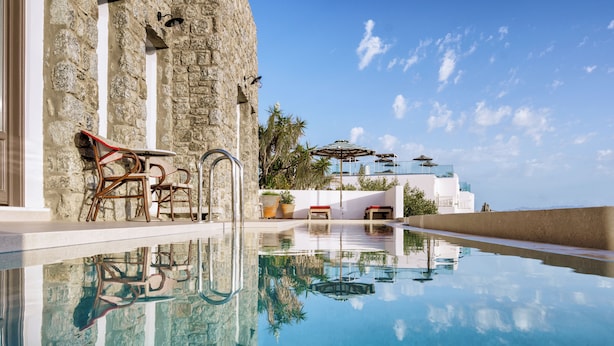 Uma piscina transparente com uma parede de pedra e um edifício branco ao fundo.
