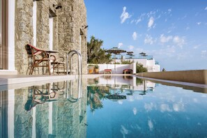 A clear swimming pool with a stone wall and a white building in the background. - Pelican Bay Hotel (Mykonos)