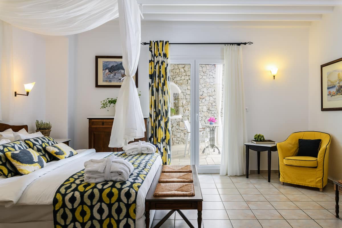 Habitación de hotel con cama, un banco, un sillón amarillo, una mesita y vistas al exterior.