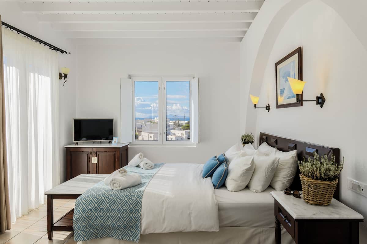Un dormitorio con cama, televisión, ventana con vistas y una lámpara fijada a la pared.