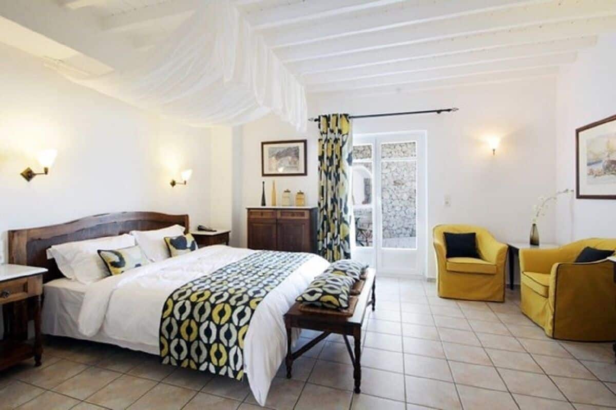Un dormitorio con cama, mesitas de noche, un banco, dos sillas, un armario y una puerta que da a un balcón.