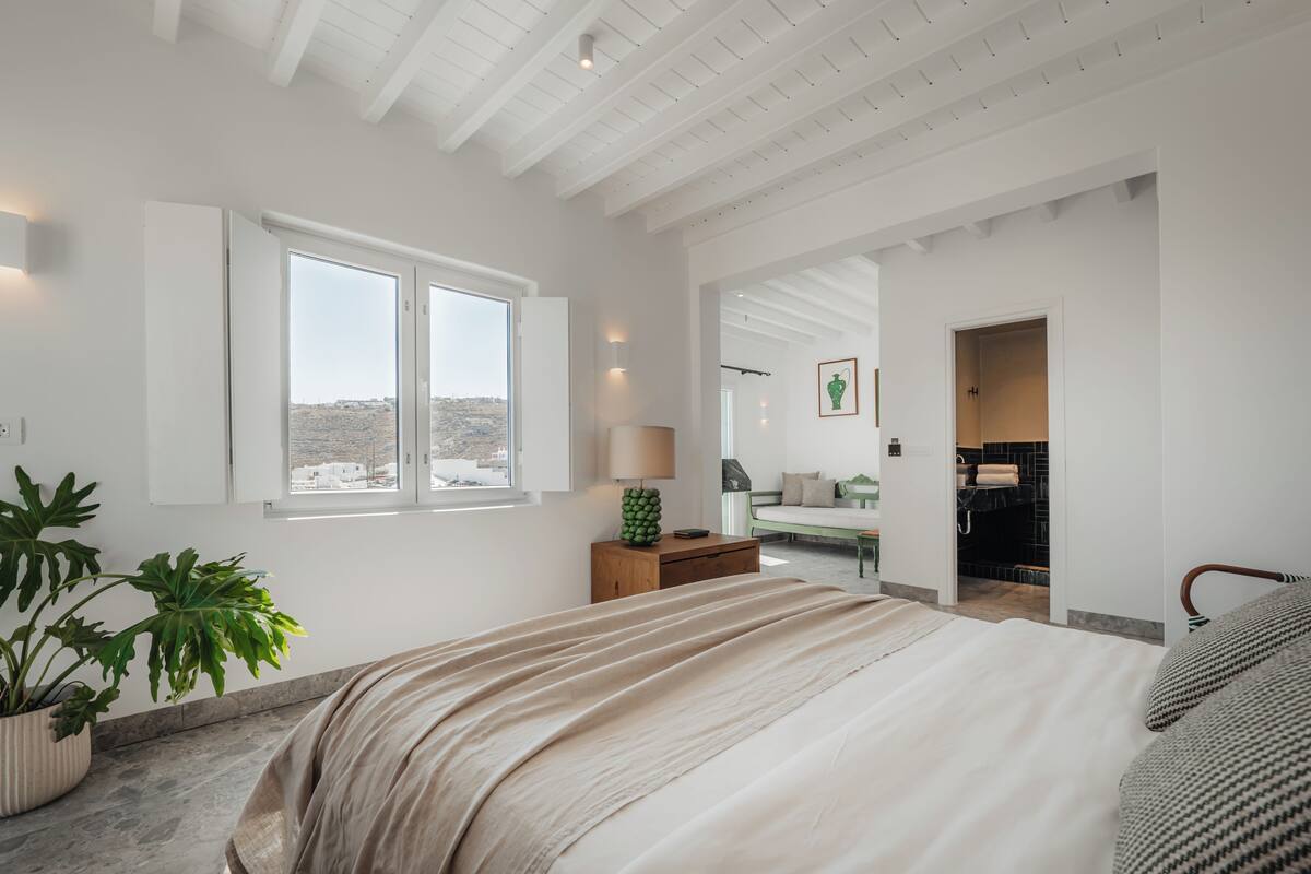 Un dormitorio amplio con una cama grande, una planta en maceta, una mesita de noche con lámpara y vistas a la ciudad desde la ventana.