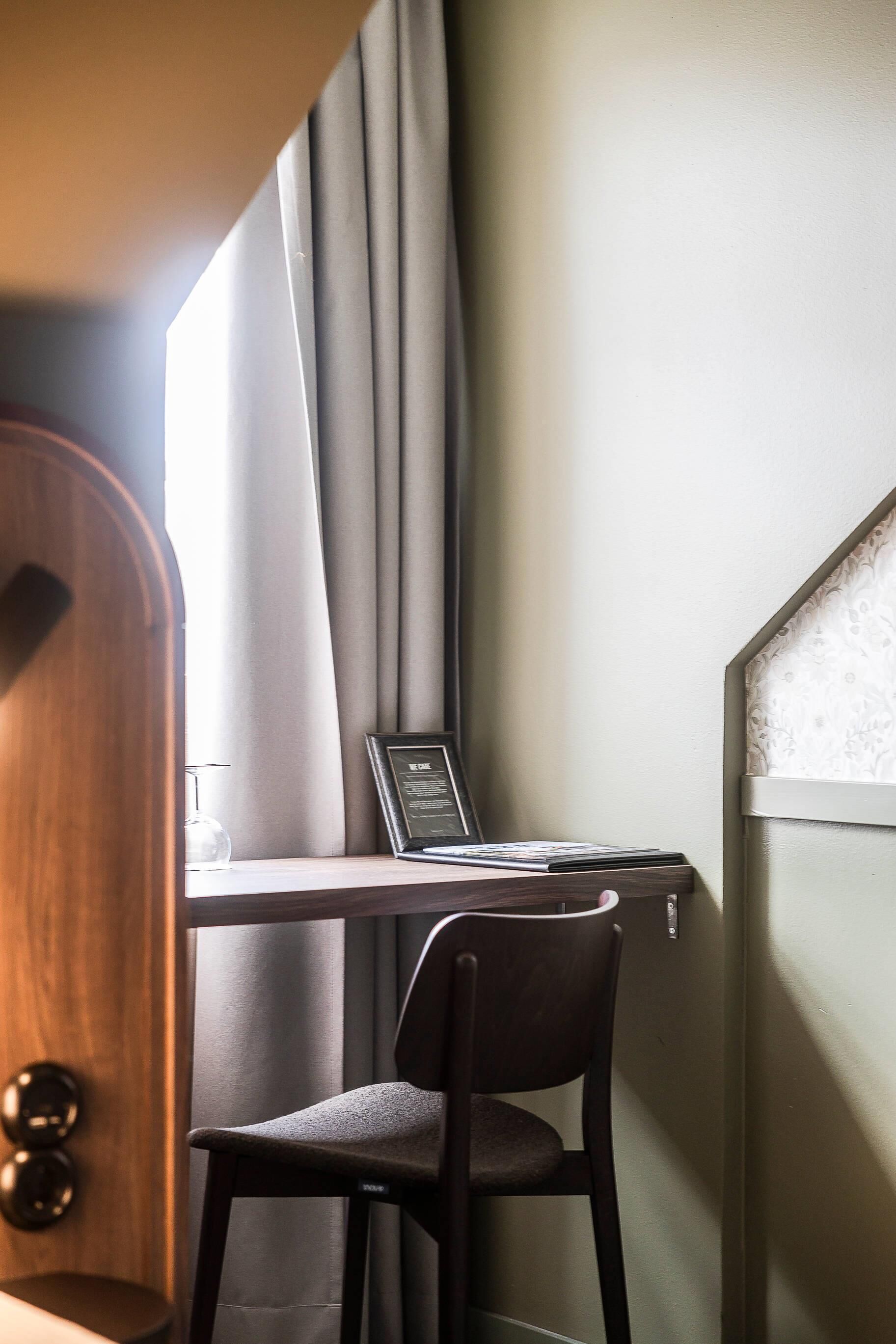 A corner of a room with a wooden door, a desk with a chair, a framed certificate, and a window with curtains.