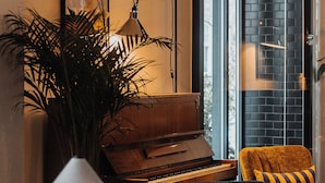 A cozy living room with a piano, a lamp, a plant, and a view of a brick wall through a glass door. - Home Hotel Grand Sundsvall (Sundsvall)