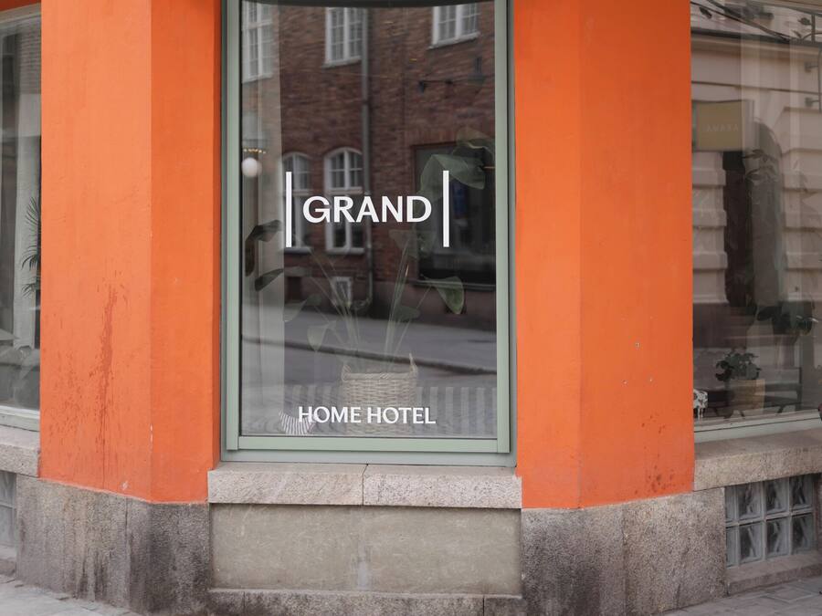 Een hotelentree met een glazen deur, een bord met de tekst 'GRAND HOME HOTEL' en een metalen luifel.