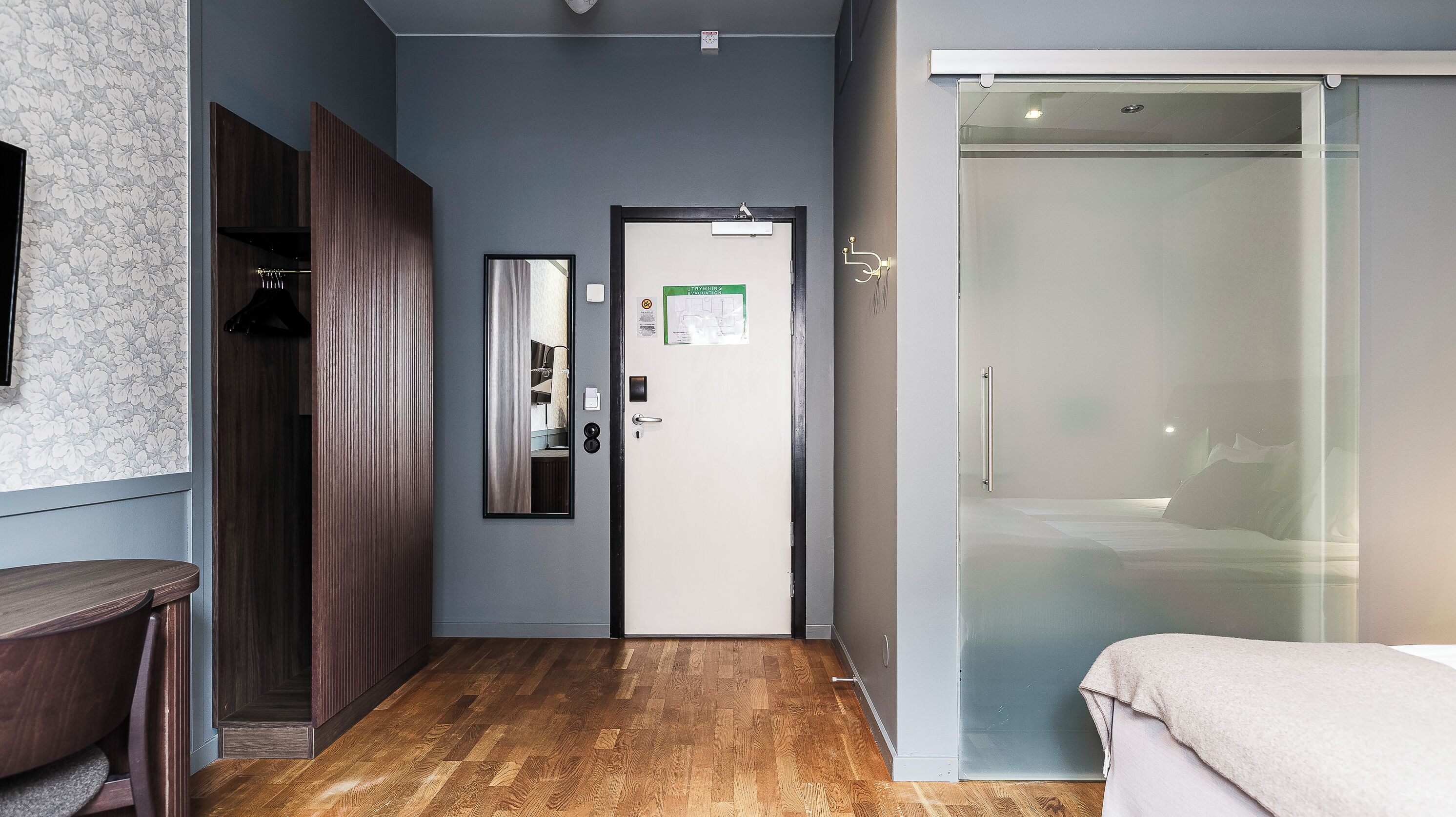 A hotel room with a wooden floor, a wardrobe, a mirror, a door with an evacuation map, and a sliding glass door leading to a balcony.
