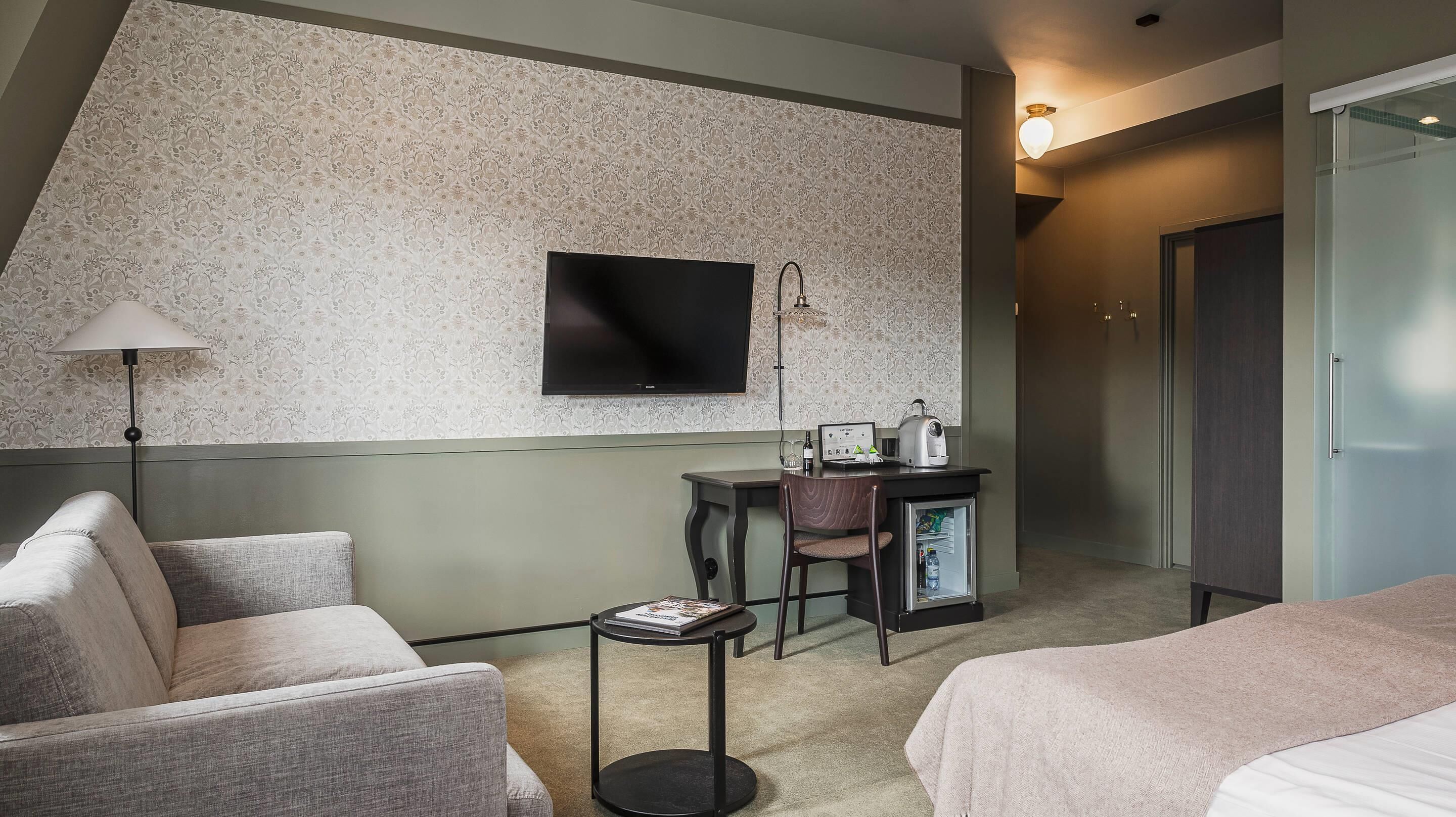 Habitación de hotel con televisión de pantalla plana en la pared, un escritorio con una computadora portátil y un hervidor, un refrigerador pequeño y dos camas.