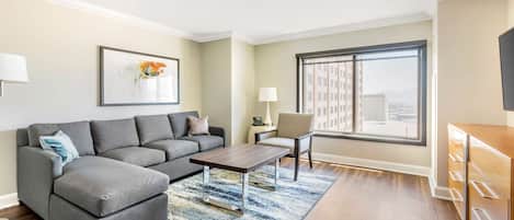 Uma sala de estar moderna com um sofá seccional cinza, uma mesa de centro de madeira e uma janela ampla com vista para a cidade.