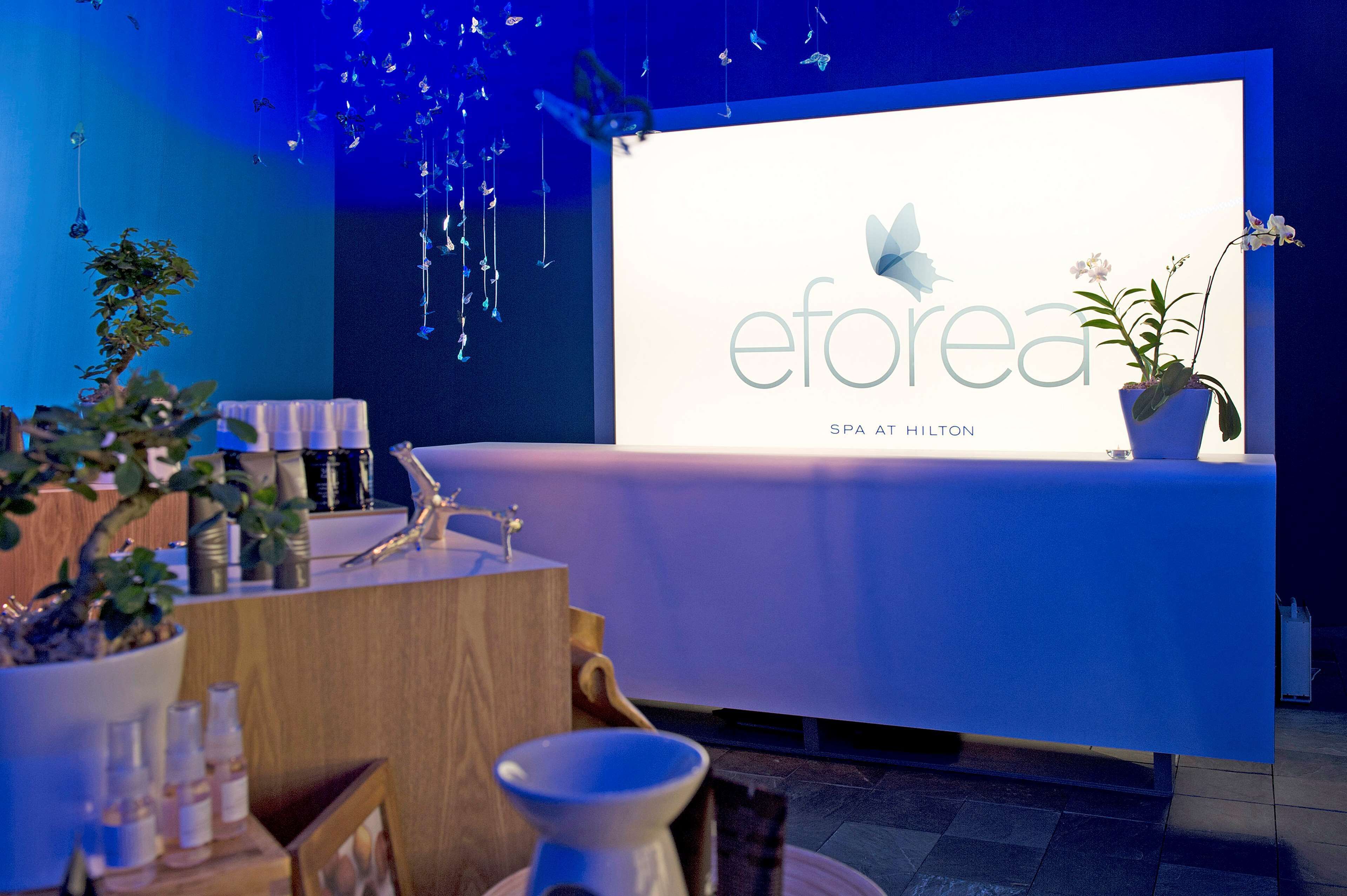 A reception area with a reception desk, a potted plant, and a wall-mounted display with the logo and name 'eForea SPA AT HILTON'.
