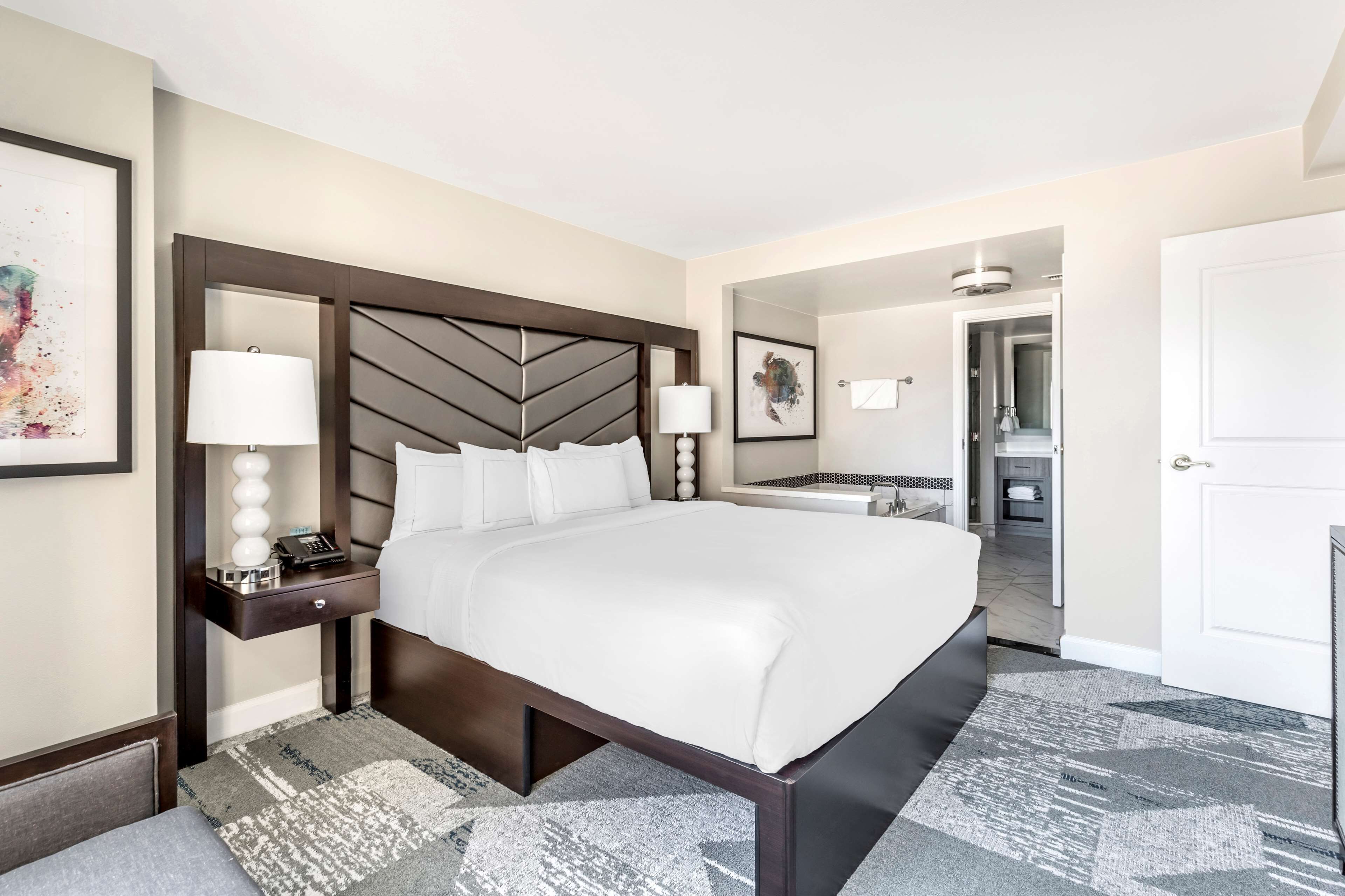 A hotel room with a large bed, a bedside table with a lamp, a nightstand, and a framed picture on the wall.