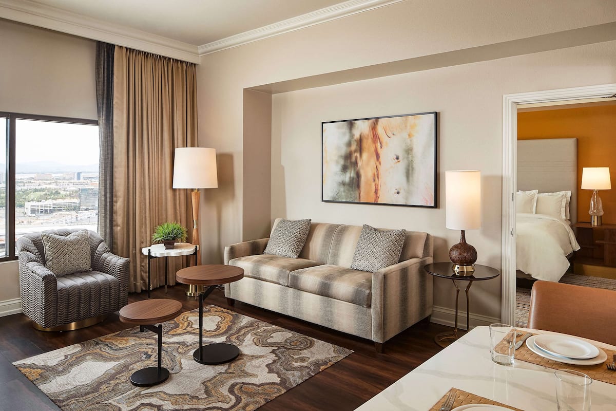 A modern hotel room with a sofa, armchair, and dining table. There is a city view through large windows, a painting on the wall, and a lamp on each side of the sofa.