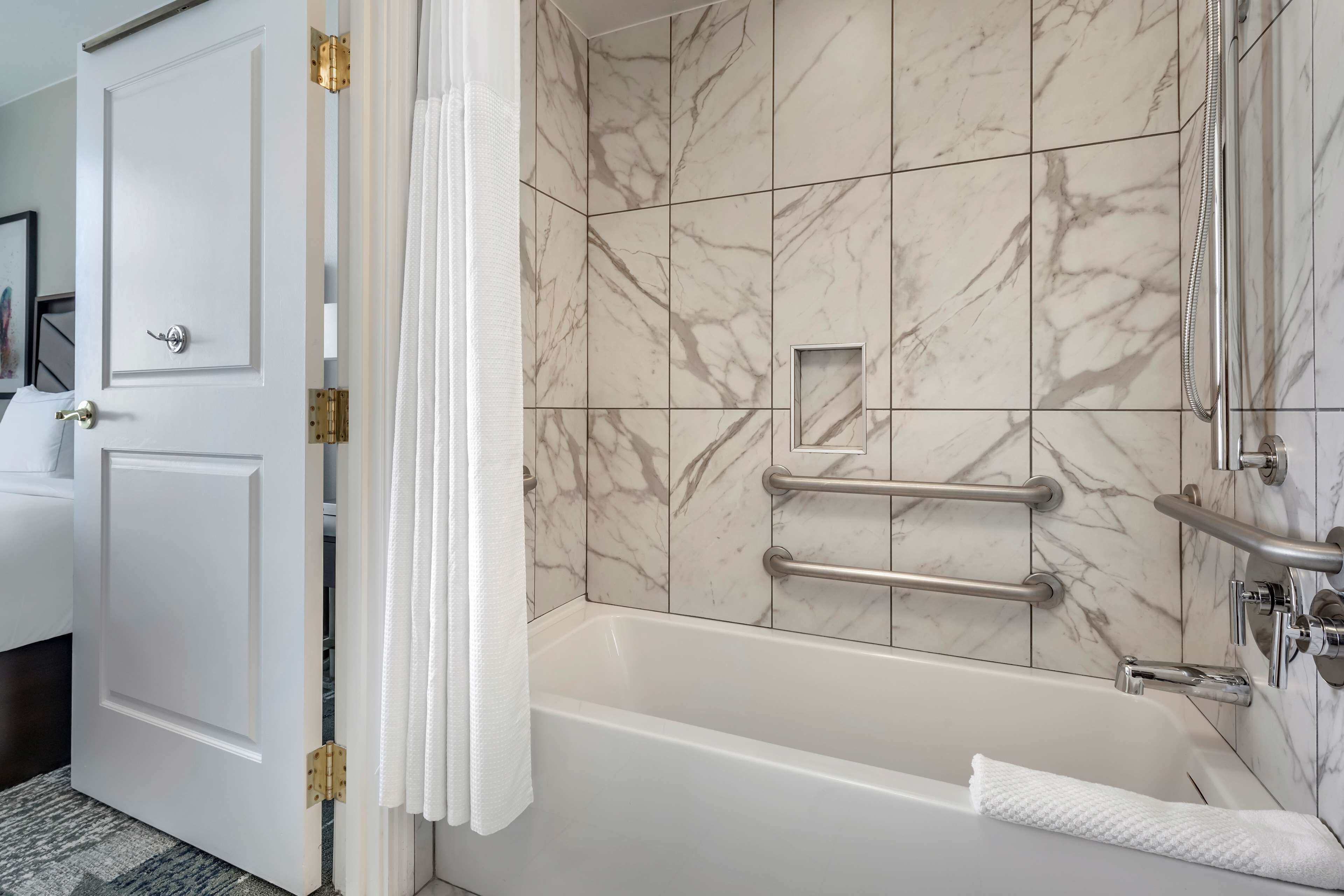A bathroom with a marble-tiled wall, a bathtub, a shower area, and a door with a towel hanging on it.