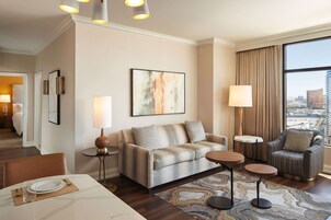 Una habitación de hotel moderna con sofá, sillón, mesa de comedor y un amplio ventanal con vista a la ciudad.