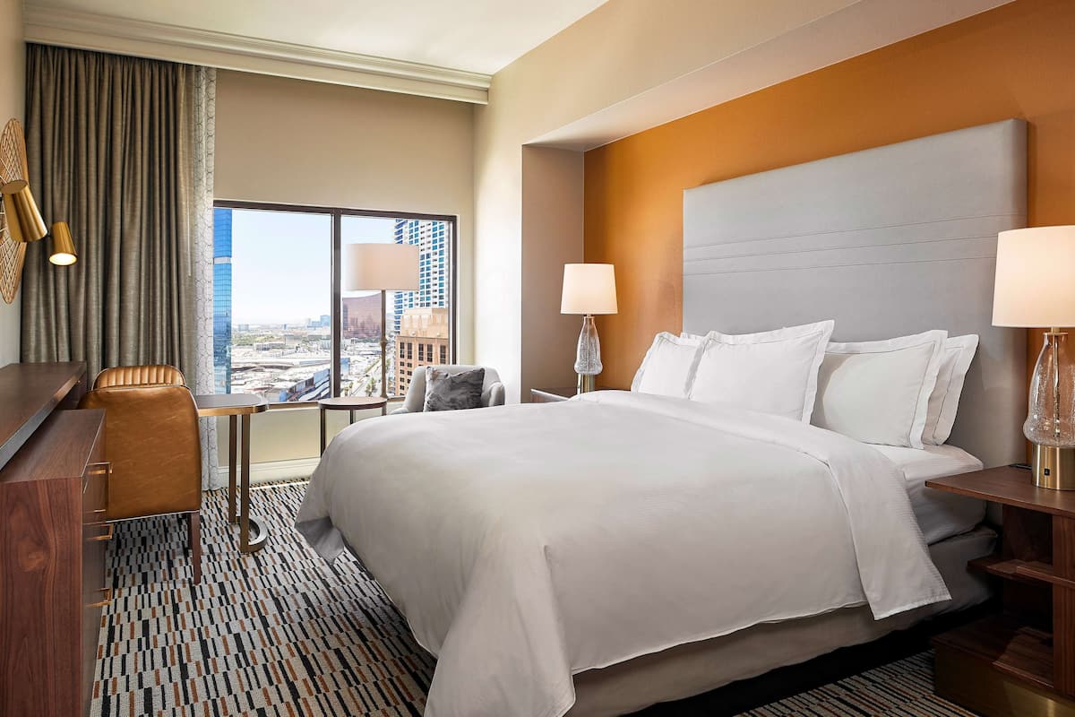 A hotel room with a large bed, two bedside tables with lamps, a desk with a chair, and a view of a cityscape through large windows.