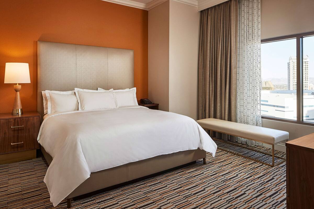 A hotel room with a large bed, a bedside table with a lamp, a bench, and a view of the city through the window.
