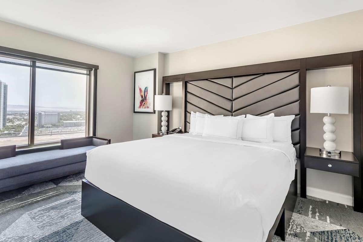 A hotel room with a large bed, a view of the city, a sofa, and a nightstand with a lamp.