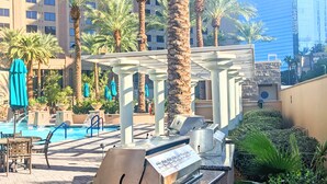 A rooftop pool area with palm trees, a barbecue grill, and a view of high-rise buildings.