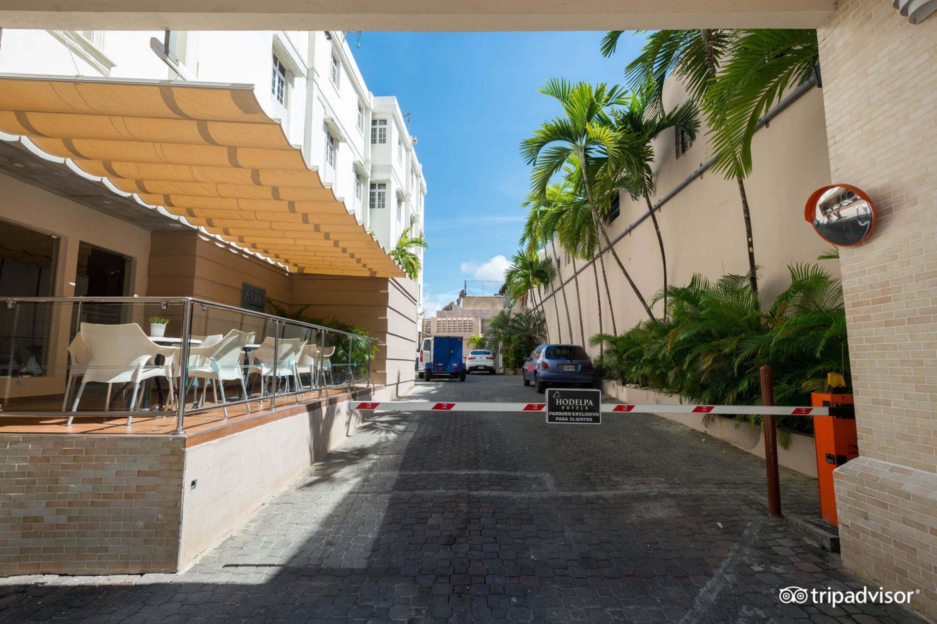Uma rua estreita com portão, palmeiras e um edifício com fachada branca e área de estar coberta ao ar livre.